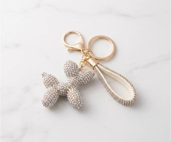 El Llavero Perrito Glam es un accesorio deslumbrante de máxima tendencia con un sofisticado acabado dorado. Destaca por su dije tridimensional en forma de perro globo, completamente cubierto de cristales iridiscentes que reflejan destellos multicolores. Equipado con mosquetón, argolla y una elegante correa brillante, combina versatilidad, lujo y un espíritu lúdico único.