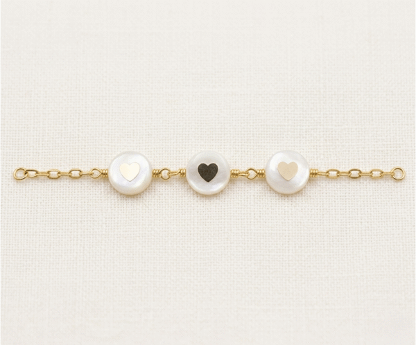 La Pulsera Aros con Corazón es una joya de elegancia atemporal que destaca por su exquisito acabado dorado. Presenta tres medallones circulares de nácar natural con delicados corazones en su centro, unidos por una refinada cadena de eslabones. Fabricada en acero quirúrgico, combina romanticismo y luz con un estilo artesanal único.