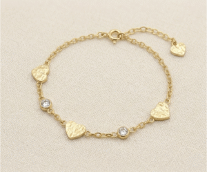 La Pulsera Corazón Colgante es una pieza de encanto artesanal y brillo sutil. Presenta delicados dijes de corazón con una textura martillada única, intercalados con cristales brillantes engastados en bisel sobre una cadena dorada. Fabricada en acero quirúrgico, esta joya combina romanticismo y modernidad, ofreciendo un estilo cálido y sofisticado que resalta en cualquier ocasión.