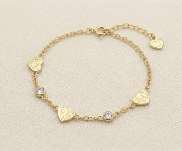 La Pulsera Corazón Colgante es una pieza de encanto artesanal y brillo sutil. Presenta delicados dijes de corazón con una textura martillada única, intercalados con cristales brillantes engastados en bisel sobre una cadena dorada. Fabricada en acero quirúrgico, esta joya combina romanticismo y modernidad, ofreciendo un estilo cálido y sofisticado que resalta en cualquier ocasión.