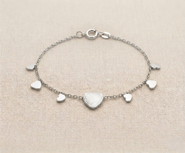 Pulsera Lluvia de Corazones _A126 La Pulsera Lluvia de Corazones es una pieza de exquisita delicadeza y encanto romántico con un acabado plateado brillante. Presenta un corazón central con textura martillada artesanal, rodeado por pequeños dijes lisos que caen en cascada sobre una fina cadena. Fabricada en acero quirúrgico, es el accesorio perfecto para un estilo femenino.